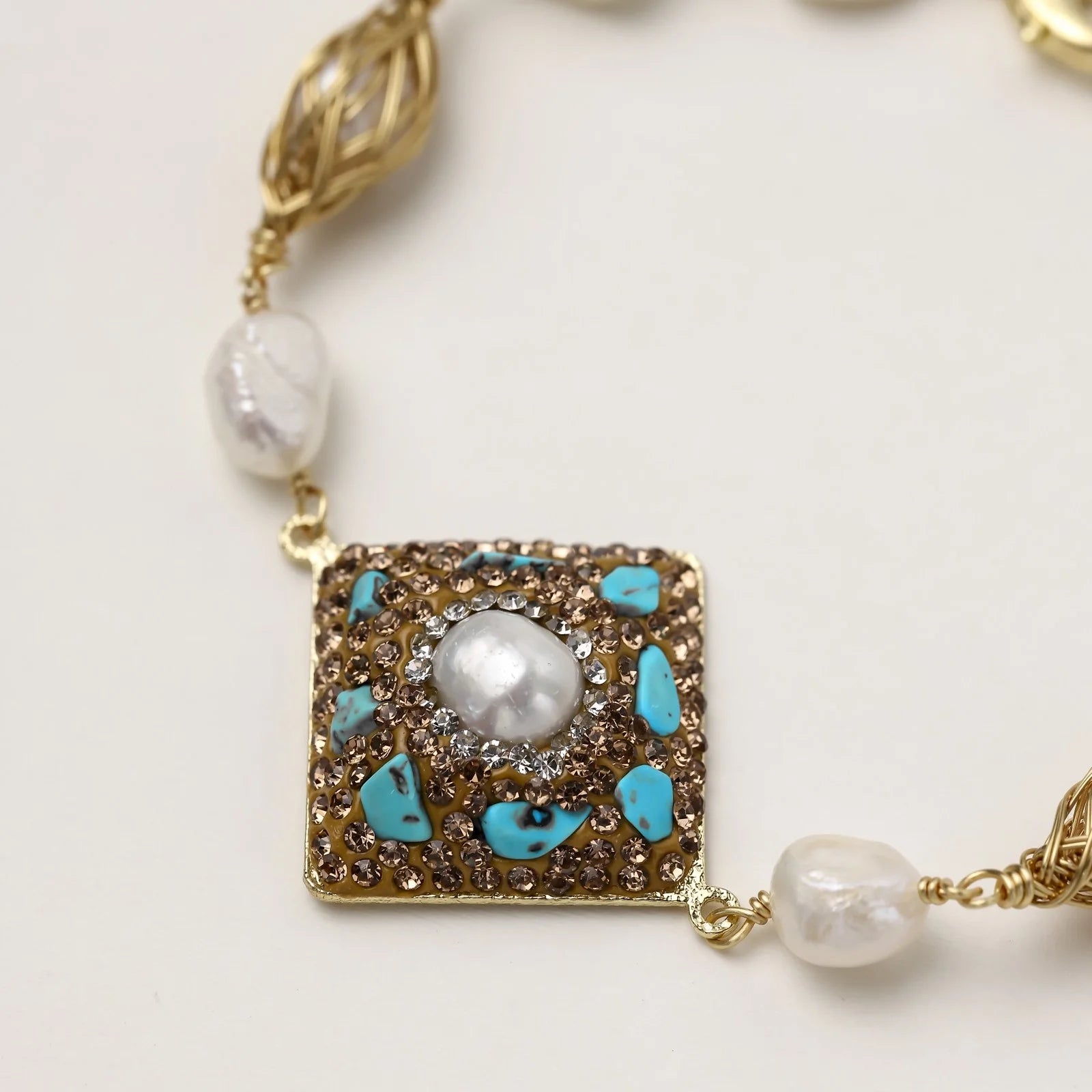 Gold-plated bracelet with baroque centerpiece, turquoise stones, freshwater pearls, and ornate chain links on cream background.