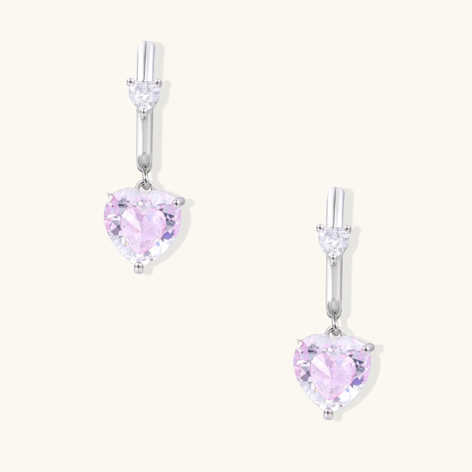 Silver huggie hoops with pink heart-cut zirconia pendants on white background, eco-friendly white gold plated brass