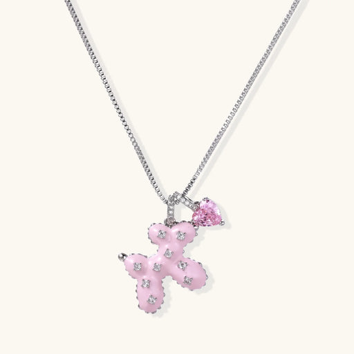 The Pink Cross Heart Necklace by Astral Signature features a pink enamel teddy bear adorned with sparkling zirconia charms, made of White Gold Plated Eco-Brass, perfect as a gift for her.