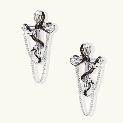 Silver dangle earrings with sculptural serpent coiled around cross shape and delicate chain accents on cream background