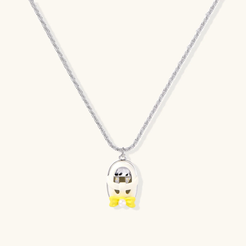 Close-up of the Cosmic Dreamer Necklace featuring a chrome egg pendant with a kawaii face and a yellow bow, designed by Astral Signature in white gold plated Eco-Brass. Perfect addition to any jewelry collection. Cosmic Dreamer Necklace, Eco-Brass Necklaces, Necklace, White Gold Plated Eco-Brass.