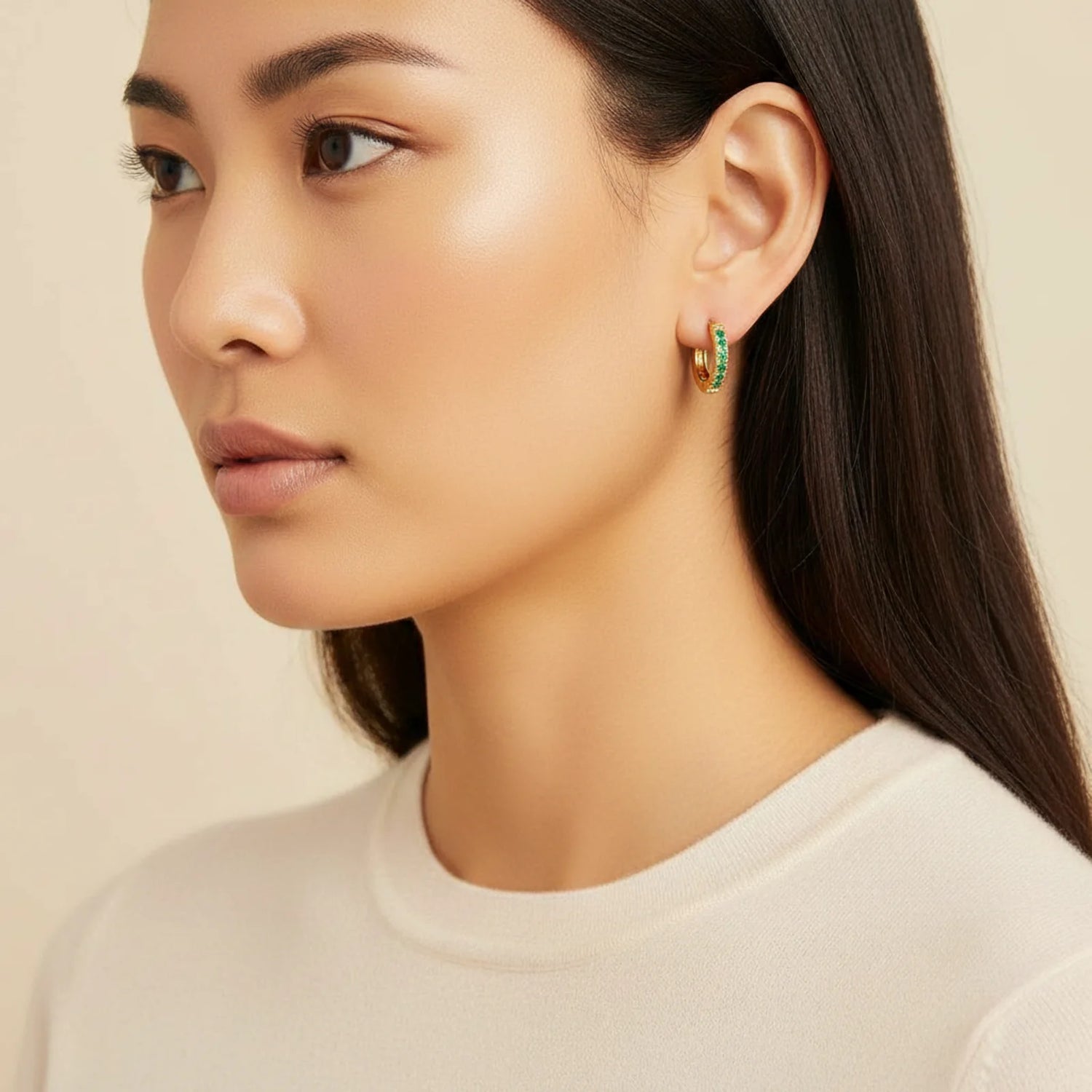 A model showcasing the Emerald Energy Huggies 18K Gold Plated by Astral Signature, featuring vibrant emerald CZ stones on gold-plated stainless steel, evoking a luxurious look. Perfect for gifting, this limited edition design is ideal for elegance in any setting. Astral Design, Astral Signature, Gift for Her, Limited Edition.