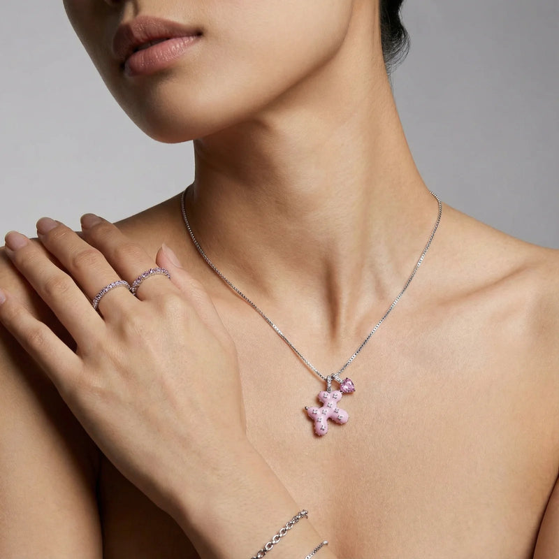 A close-up of a model wearing the Pink Cross Heart Necklace by Astral Signature, displaying the charming pink teddy bear design against her neckline, made from White Gold Plated Eco-Brass with zirconia.