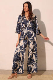 A model showcasing the Astral Women's Jumpsuit - Navy Orchid Lily Satin Cape by TYCHE, featuring a flowing design and a chic abstract floral print. Perfect for weddings and parties, this elegant jumpsuit embodies modern grace. APPAREL, CCPRODUCTS, JUMPSUITS & ROMPERS, Navy, NEW ARRIVALS, Women's, Women's Clothing, Women's Fashion, Women's Jumpsuit, Women's Style.
