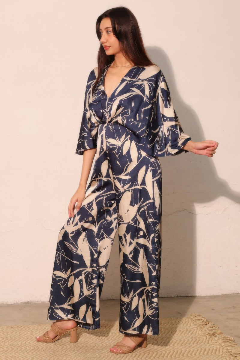 A stylish display of the Astral Women's Jumpsuit - Navy Orchid Lily Satin Cape by TYCHE, captured from a side angle revealing its fluid cape movement and elegant silhouette. A perfect addition to any woman's wardrobe for formal gatherings. APPAREL, CCPRODUCTS, JUMPSUITS & ROMPERS, Navy, NEW ARRIVALS, Women's, Women's Clothing, Women's Fashion, Women's Jumpsuit, Women's Style.