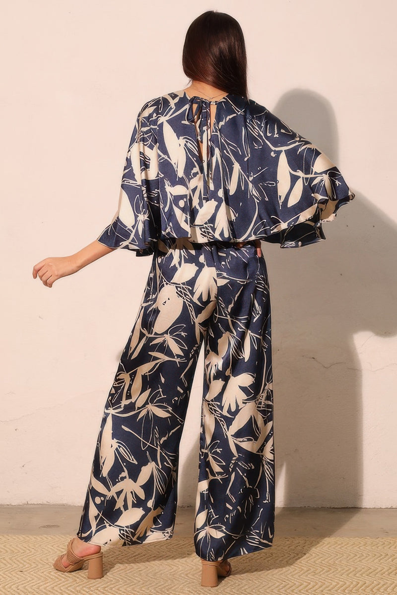 Rear view of the Astral Women's Jumpsuit - Navy Orchid Lily Satin Cape by TYCHE, highlighting its stylish cape overlay and botanical print. Ideal for elevated occasions, this jumpsuit redefines women's elegance. APPAREL, CCPRODUCTS, JUMPSUITS & ROMPERS, Navy, NEW ARRIVALS, Women's, Women's Clothing, Women's Fashion, Women's Jumpsuit, Women's Style.
