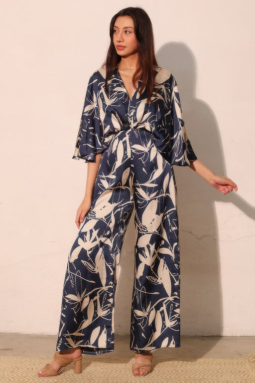 A model showcasing the Astral Women's Jumpsuit - Navy Orchid Lily Satin Cape by TYCHE, featuring a flowing design and a chic abstract floral print. Perfect for weddings and parties, this elegant jumpsuit embodies modern grace. APPAREL, CCPRODUCTS, JUMPSUITS & ROMPERS, Navy, NEW ARRIVALS, Women's, Women's Clothing, Women's Fashion, Women's Jumpsuit, Women's Style.