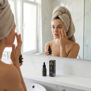 A woman applying Caffeine Gel Booster - Energizing Formula for Puffiness by Astral Signature in front of a mirror. The amber packaging highlights its anti-age and hydrating properties, targeting under-eye fatigue and damaged skin barriers. Ideal for oily and mature skin types, this natural certified booster features caffeine as a key active ingredient, providing a refreshing, oil-free face-care solution.