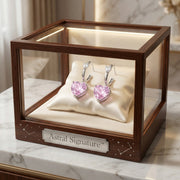Pink Heart Drop Hoops by Astral Signature elegantly displayed in a glass case, showcasing their exquisite design with pink zirconia. These Eco-Brass earrings make the ideal gift for her, featuring durable White Gold Plated Eco-Brass.